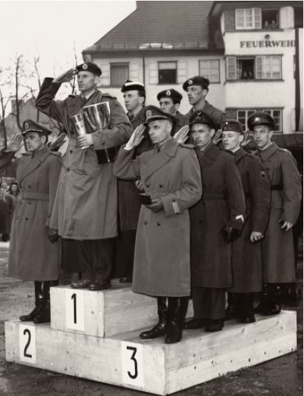 Three four-man teams stand at attention on a medal podium while saluting in military boots, overcoats, berets, and peaked caps.